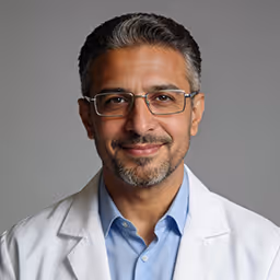 Smiling middle-aged man with glasses, salt-and-pepper hair, and beard wearing a blue shirt and white lab coat against a gray background.