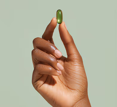 Hand with dark skin holding a green translucent capsule pill between thumb and index finger against a light green background.